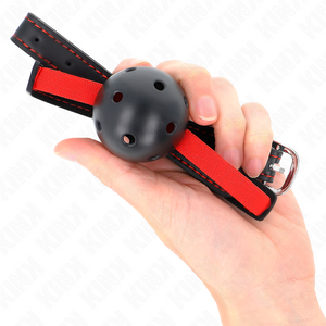 Bâillon à boule noire perforée posé dans une main avec la sangle rouge et noire